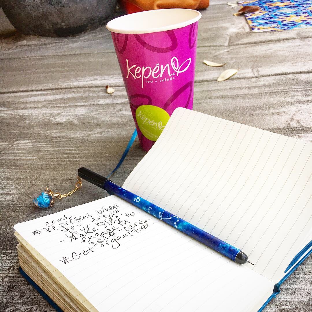An open journal with a pen sitting on the spine with a pink paper cup labeled Kepen